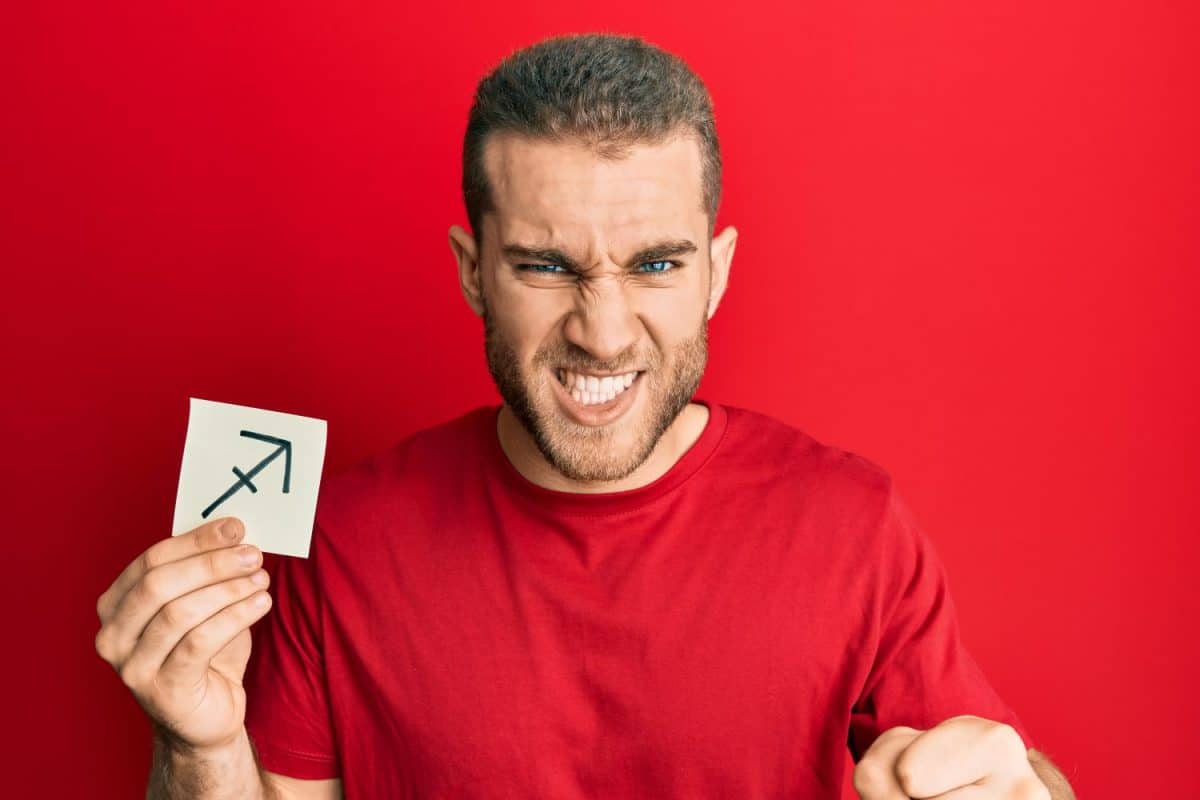 Young caucasian man holding paper with sagittarius zodiac sign annoyed and frustrated shouting with anger, yelling crazy with anger and hand raised. Sagittarius Zodiac: Personality Traits, Sign, and More