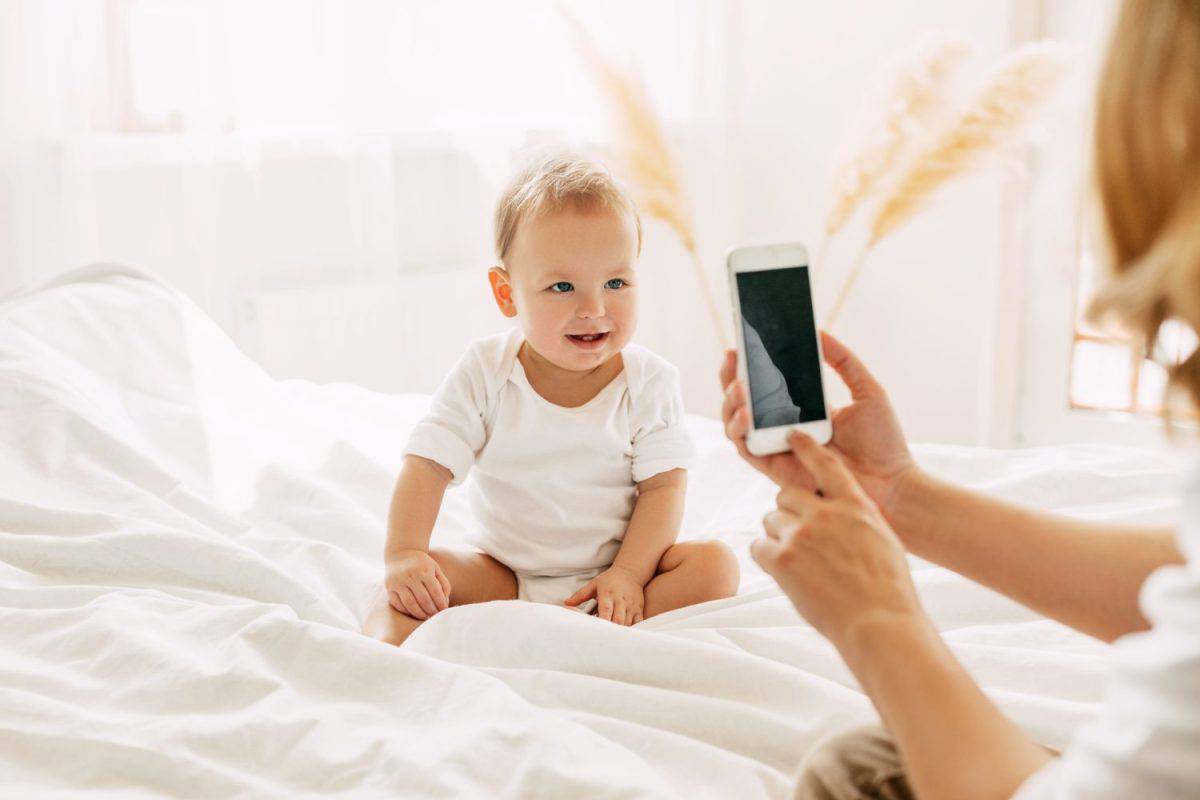 Cute baby is sitting on the bed and posing for his mom, mom is taking pictures of her baby on her mobile phone. Modern technologies, space for text