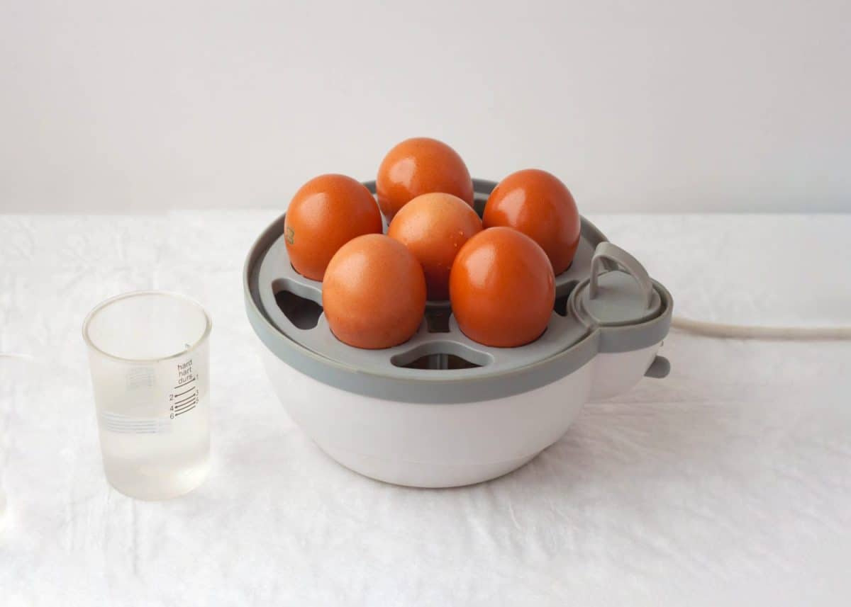 Six chicken eggs are in an egg cooker, there is a glass of water next to it.