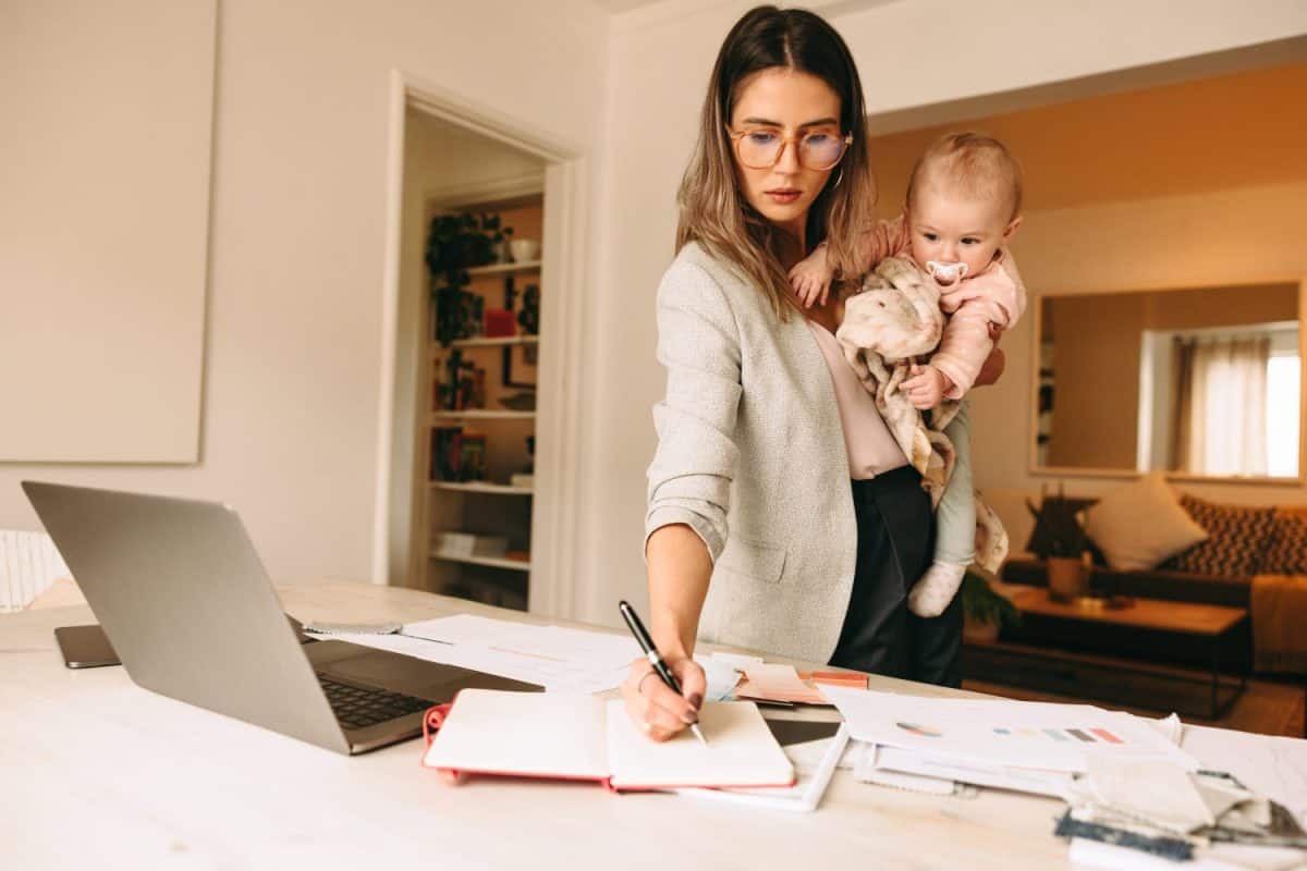 Female interior designer making notes while holding her baby. Multitasking mom planning a new project in her home office. Creative businesswoman balancing work and motherhood.