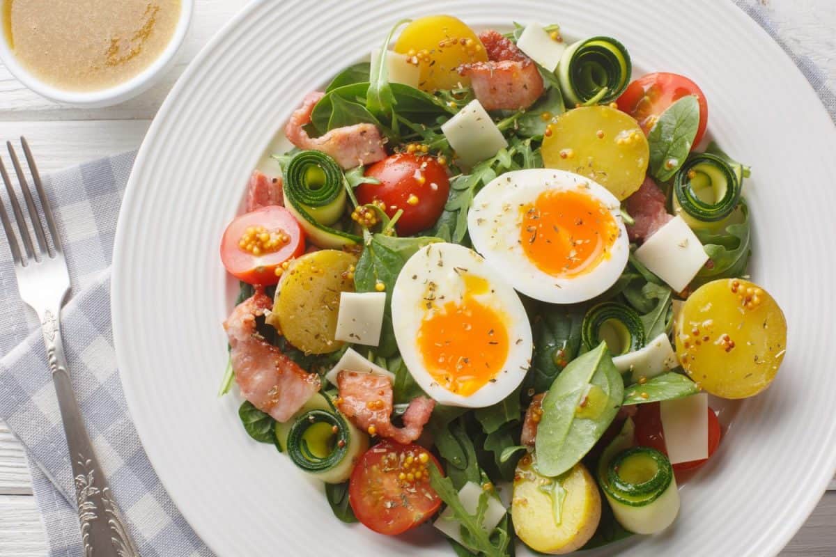 Homemade salad with vegetables, bacon, cheese and soft-boiled eggs seasoned with vinaigrette sauce close-up on a plate on the table. Horizontal top view from above