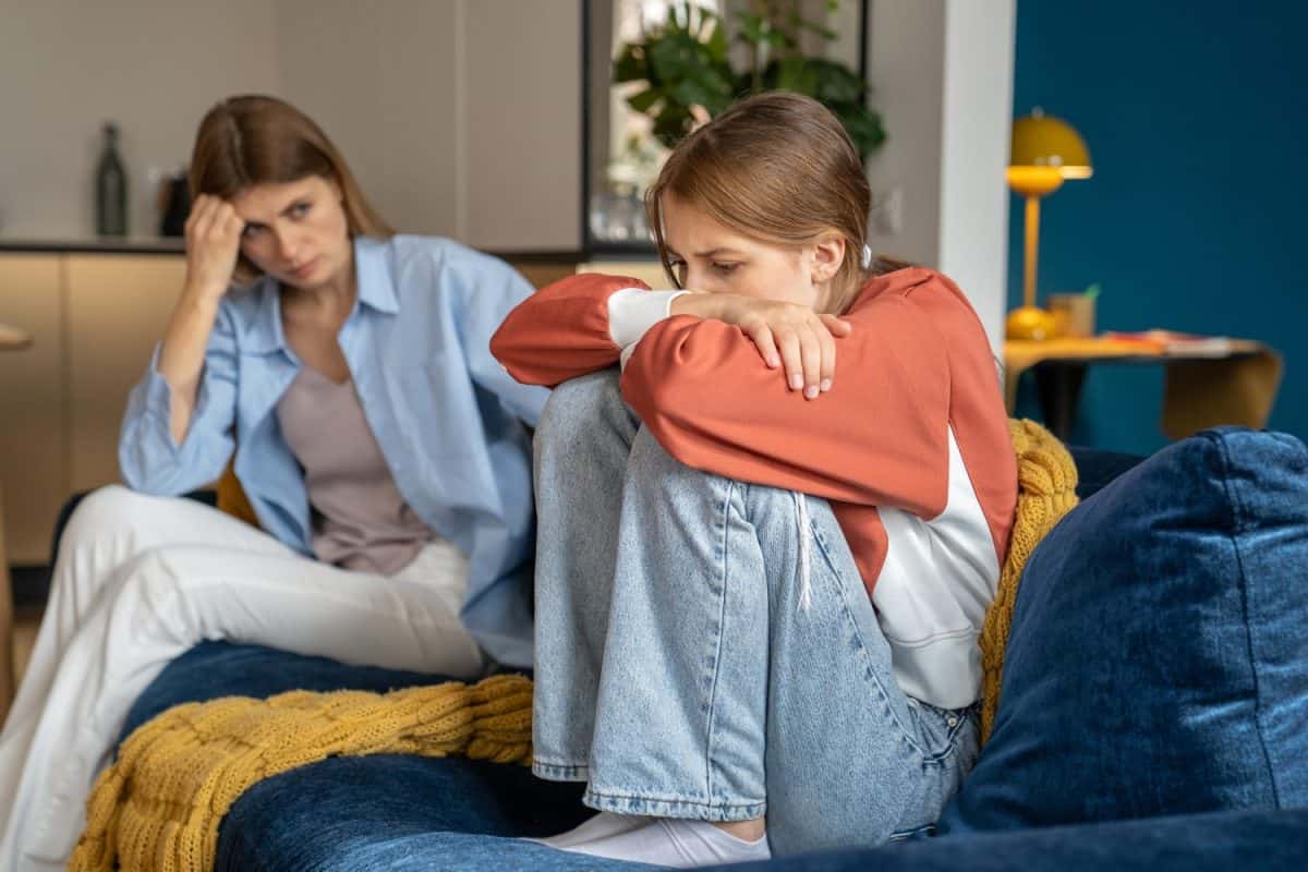 Upset worried mother looking at crying moody teenage daughter, sitting together on sofa, upset frustrated mom and teen girl child not talking after argument, having communication problems