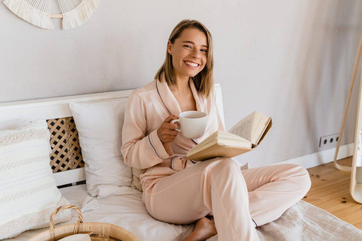 pretty smiling woman relaxing at home on bed in morning in pajamas reading book drinking coffee or tea