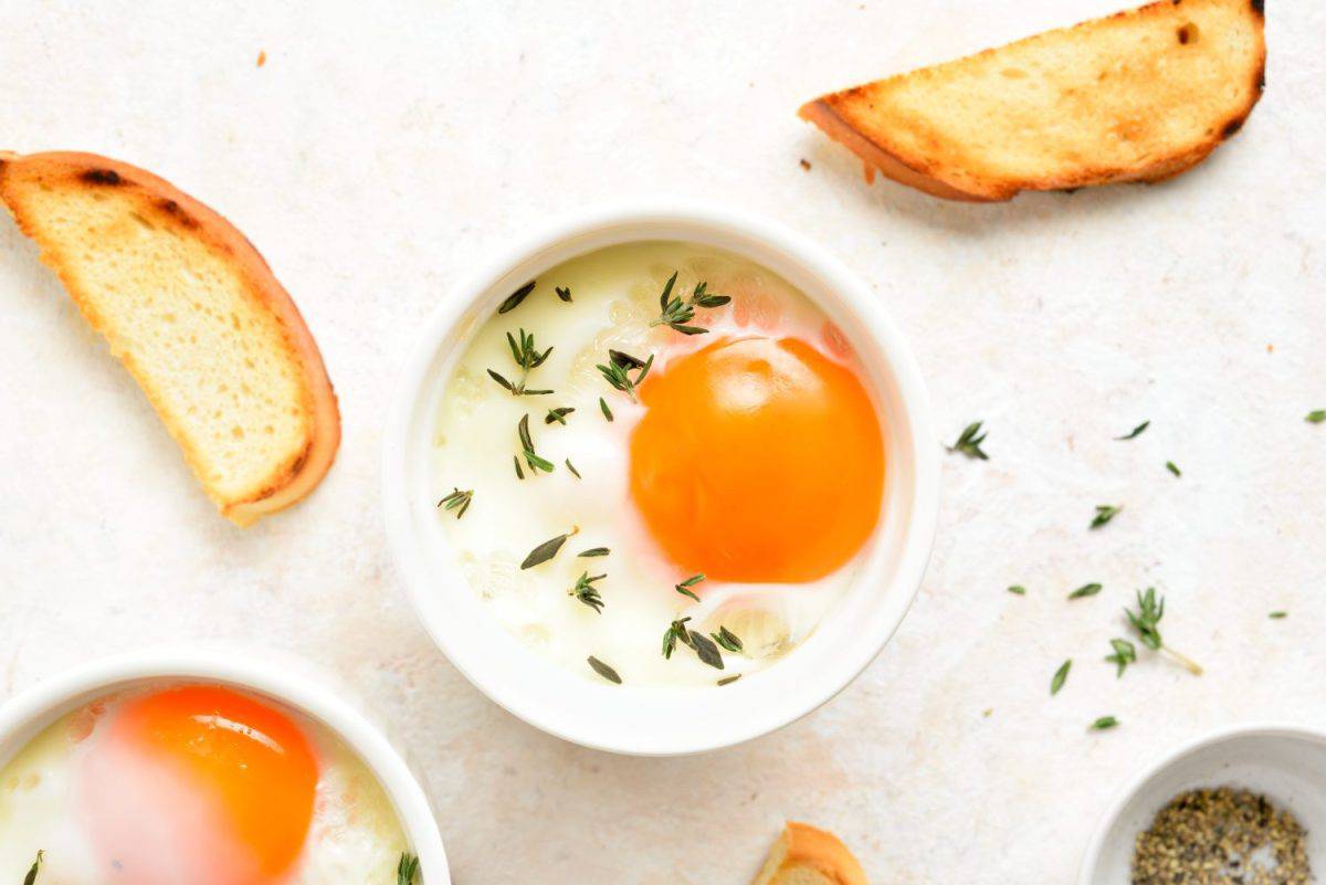 Eggs en cocotte (baked eggs) with cream, сheese and herbs served with toasted bread for dipping. Light stone background, top view, flat lay