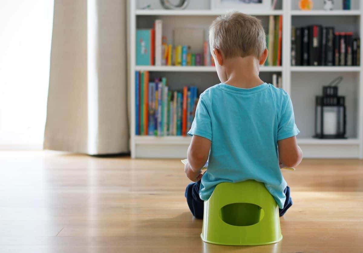 Rear view of a 3-year-old Caucasian boy in light blue t-shirt sitting on green potty reading book, large bookcase full of books in background. Potty training concept. Love reading concept. Copy space