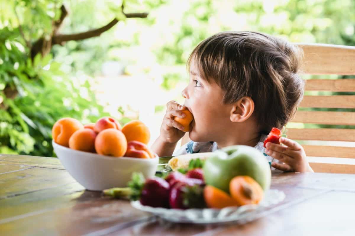 Young boy enjoying fresh fruit at a table in the garden, surrounded by vibrant greenery and various fruits.