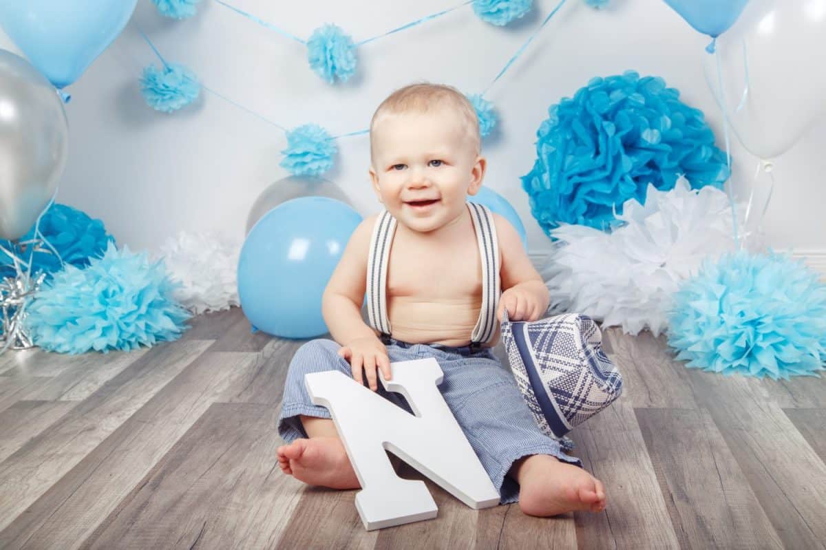 Portrait of cute adorable Caucasian baby boy with blue eyes barefoot in pants with suspenders and hat, sitting on wooden floor in studio, holding large letter N, looking away, first year concept