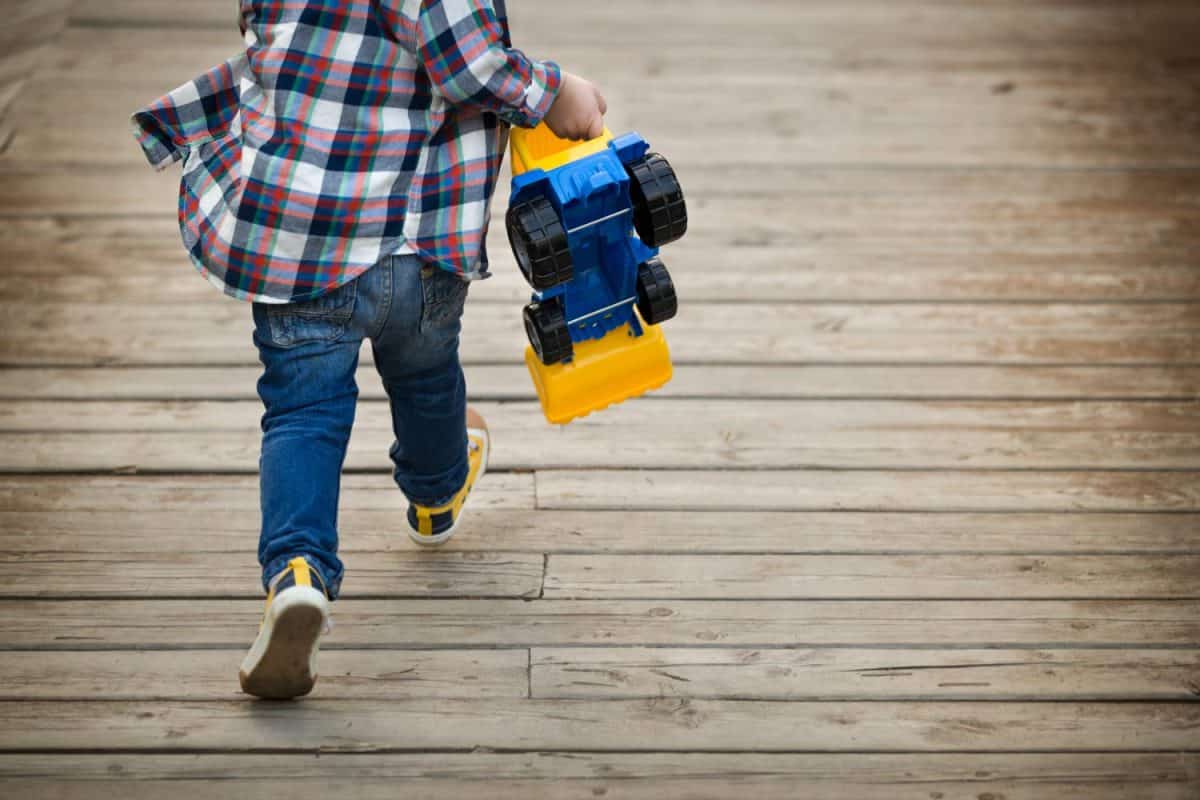 Back view on little toddler boy with his toy tractor in his hands running on wooden bridge outdoors. Child rushing away. Lifestyle