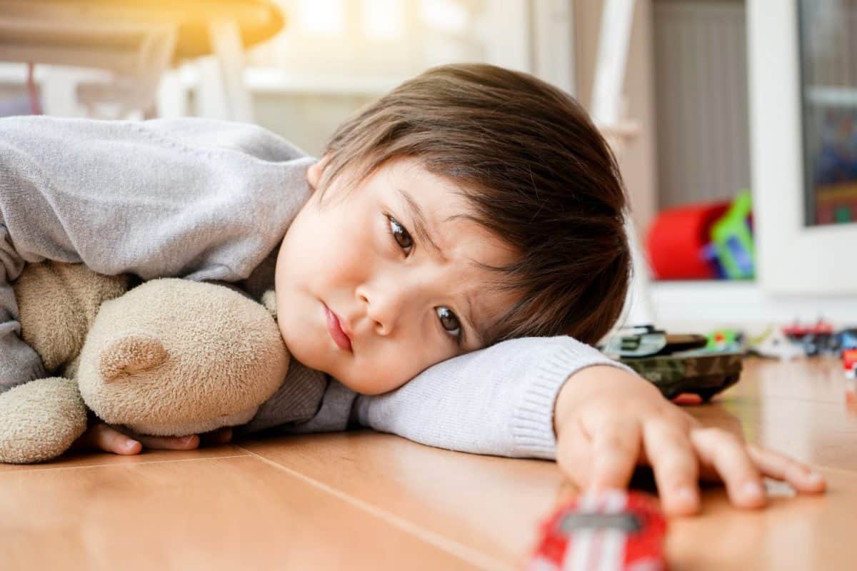 Unhappy face of Toddler boy lying down on wooden floor with his teddy bear at home