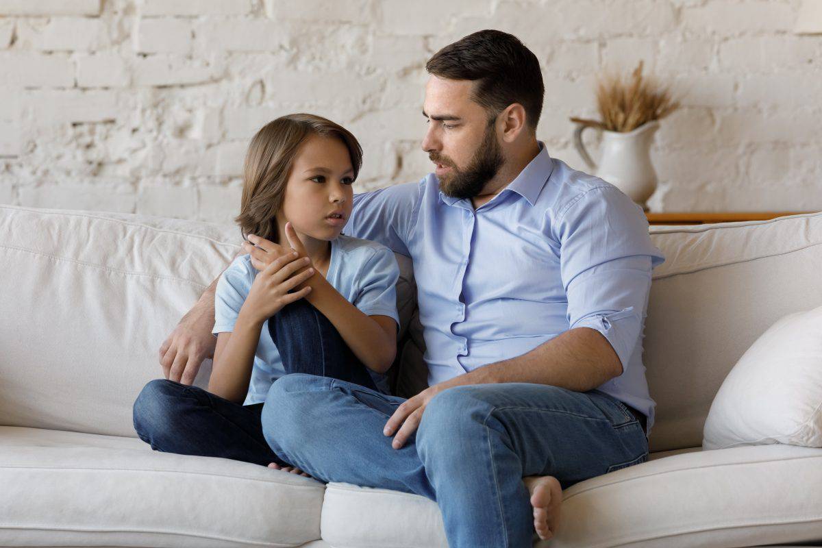 Serious father listen to his pre-teen little son talking seated on sofa at home, speaking spend time together at home. Cute boy share problems, ask advice to dad. Communication, care and trust concept