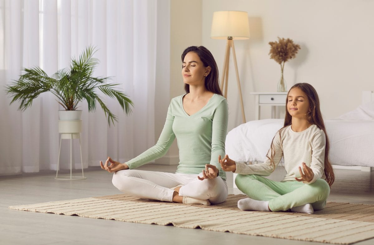 Family yoga. Beautiful young woman and her charming little daughter are smiling while doing yoga together at home. Family sits in lotus position on floor in living room. Mom teaches child to meditate.