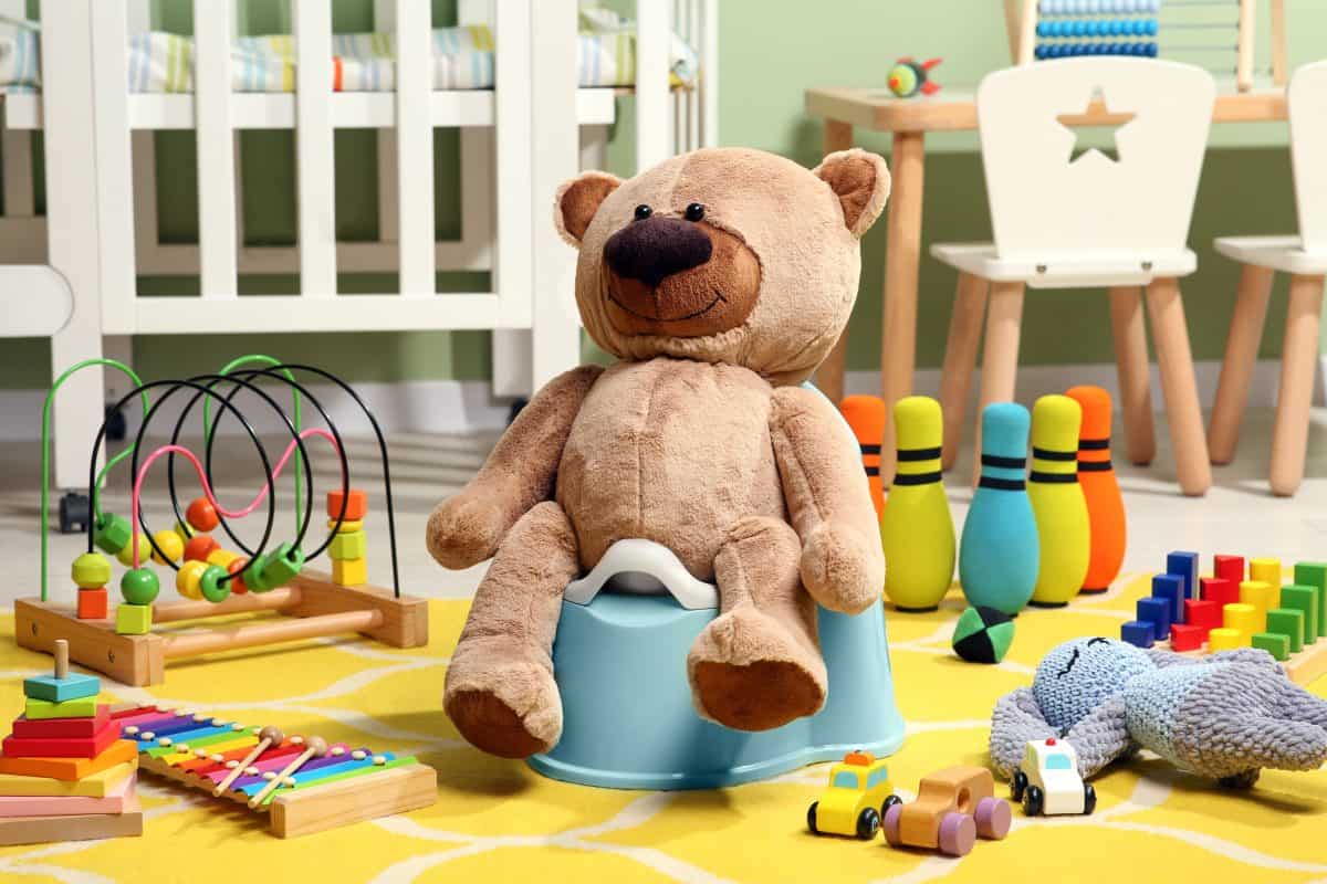 Teddy bear on light blue baby potty and many other toys in room. Toilet training