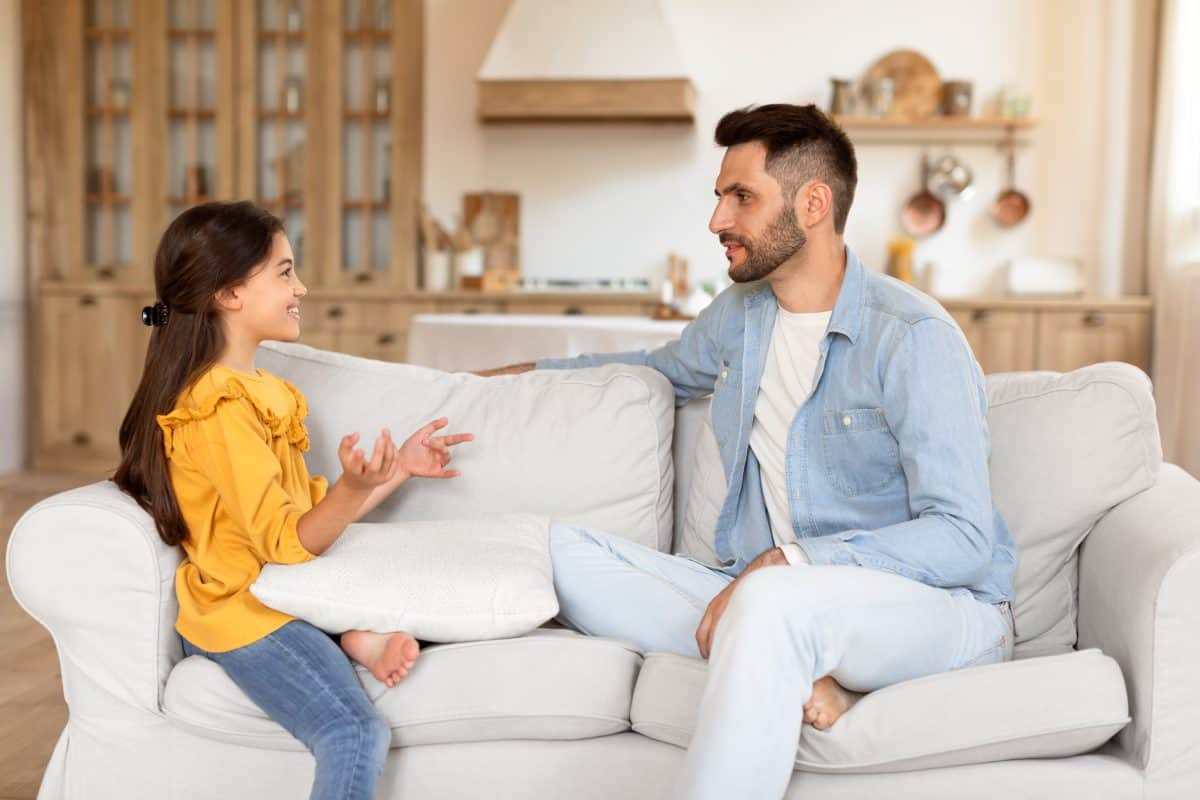 Arabic Preteen Daughter Smiling And Talking To Dad, Sharing Fun Moment Of Her Day, Bonding And Spending Time Together On Sofa At Home. Father Listens To Kid's Stories On Weekend