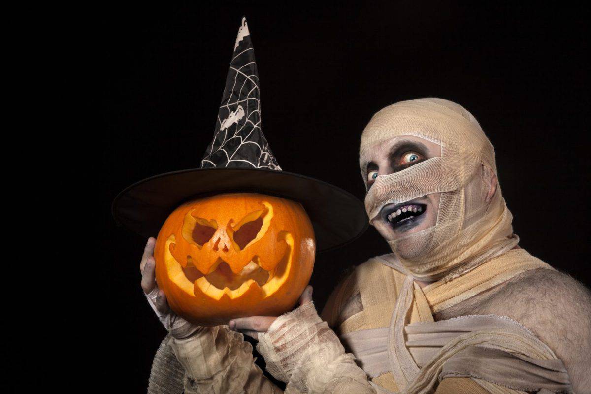 Scary Mummy holding Halloween pumpkin . Funny smiling young man in mummy costume with holyday pumpkin