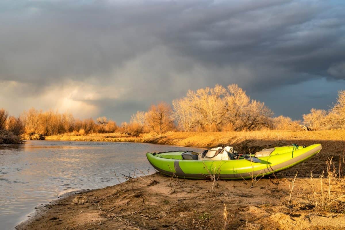 whitewater inflatable kayak with a paddle and waterproof duffel on a river shore - St Vrain Creek near Platteville, Colorado in early spring scenery with storm clouds at sunset