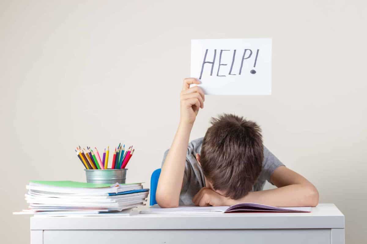 Sad tired frustrated boy sitting at the table with many books and holding paper with word Help. Learning difficulties, education concept.