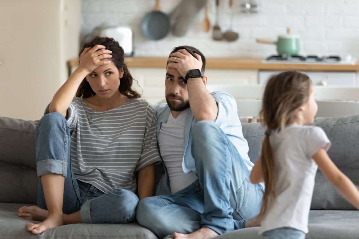 Tired mother and father having problem with noisy naughty daughter shouting and running around sofa at home, exhausted parents touching foreheads, sitting on couch, too active kid concept