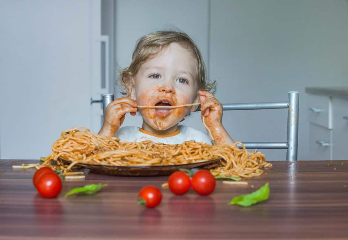 Funny baby child getting messy eating spaghetti with tomato sauce from a large plate, by itself with his hands, at home. Why servers cringe when families with small kids are seated in their section