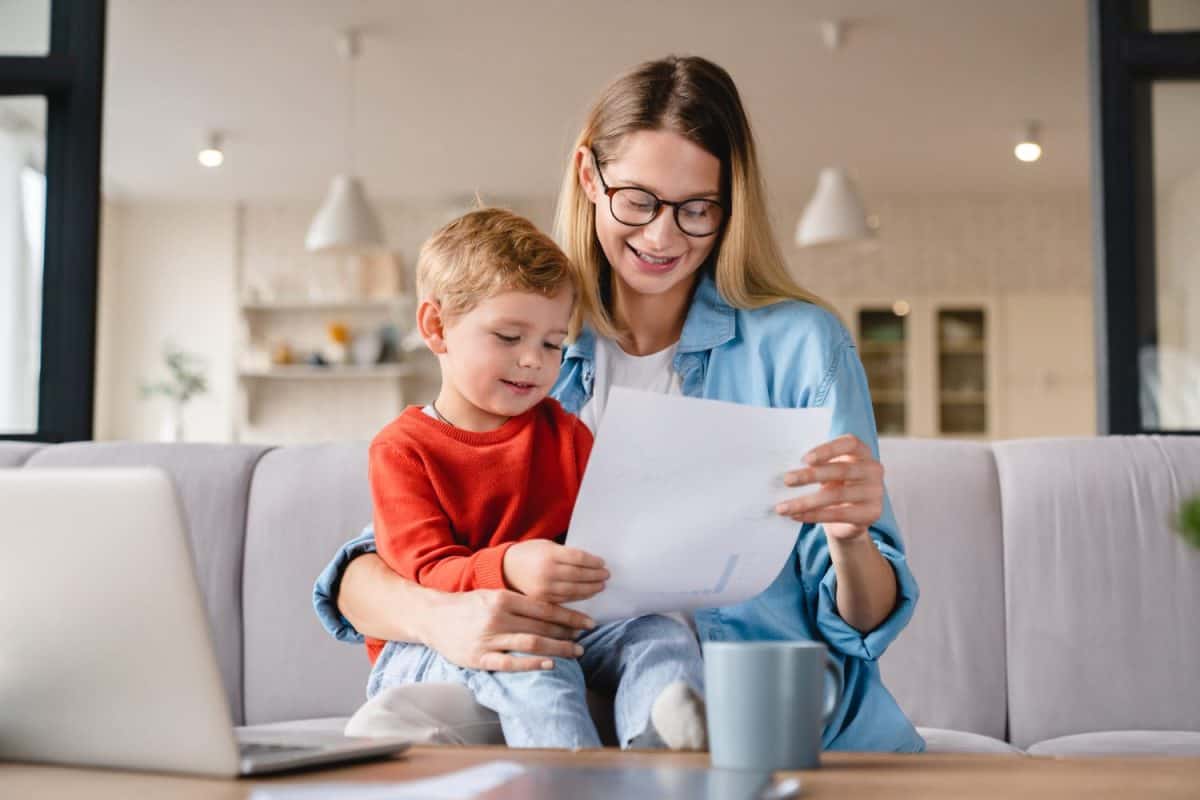 Little small toddler kid child son helping her busy young mother with freelance remote work holding papers, documents and domestic bills at home on lockdown during maternity leave