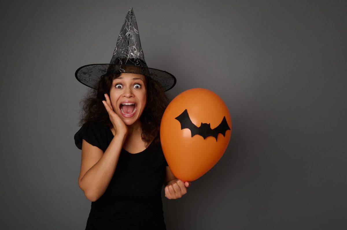 Scared woman in carnival witch costume and wizard hat, screams, holds an orange colorful balloon and puts her hand on her cheek, looking at camera. Halloween concept on gray background with copy space