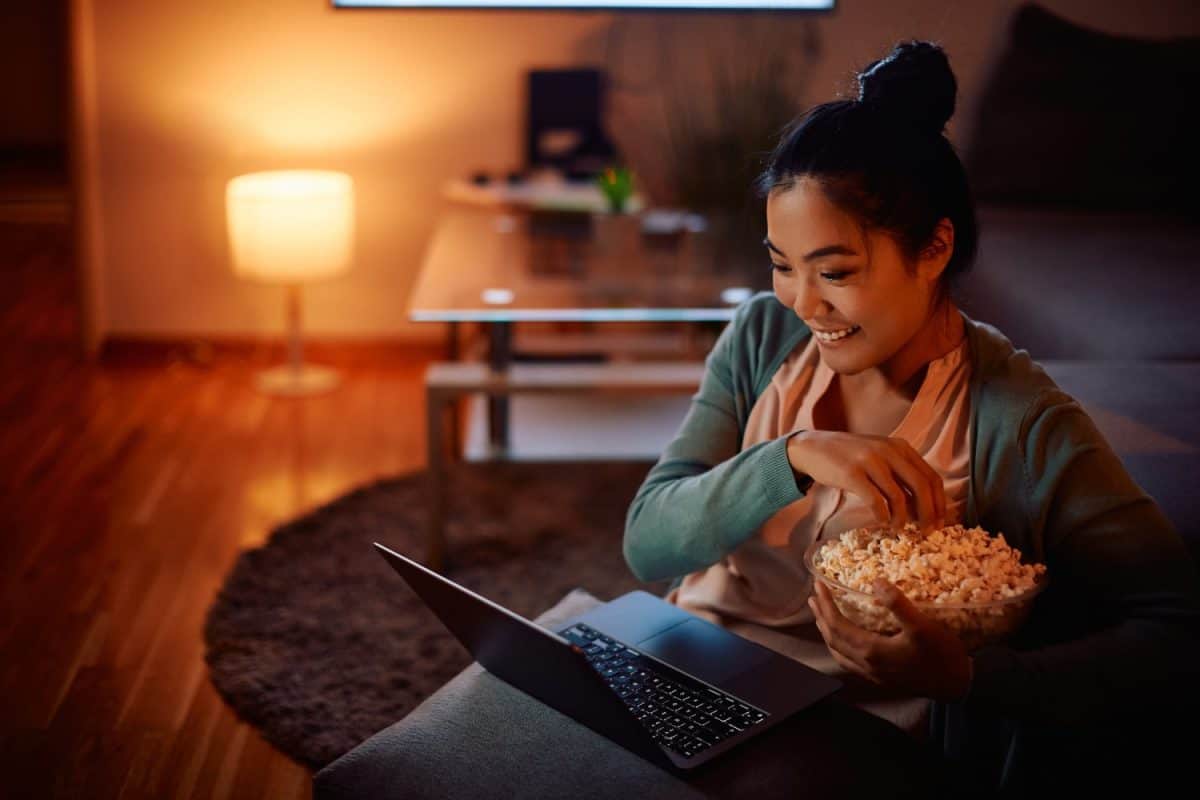 Happy Asian woman eating popcorn while watching her favorite series online on laptop in the evening at home.