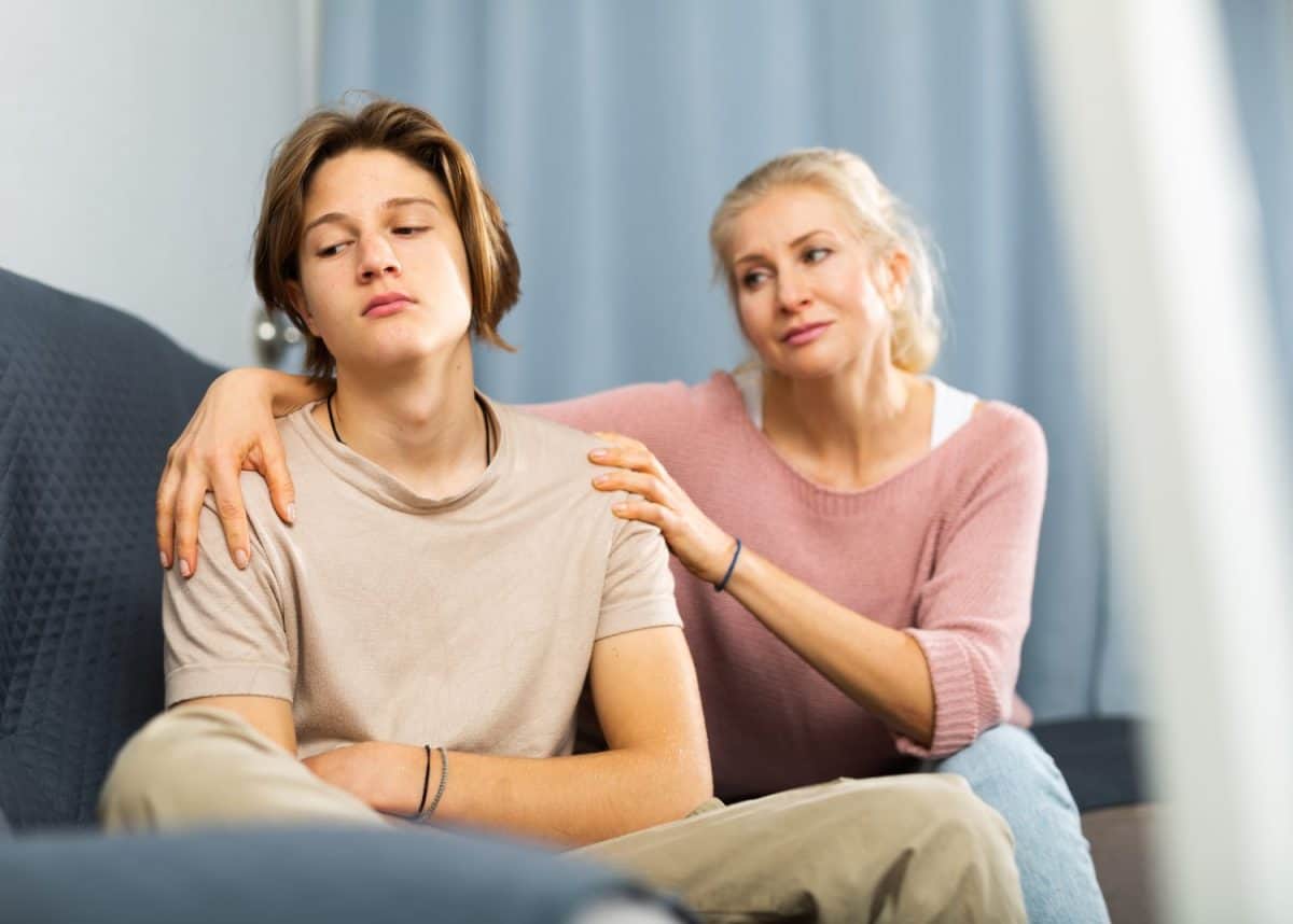 Portrait of upset and offended teenager sitting on sofa while mother soothing him. Mood swings and puberty concept