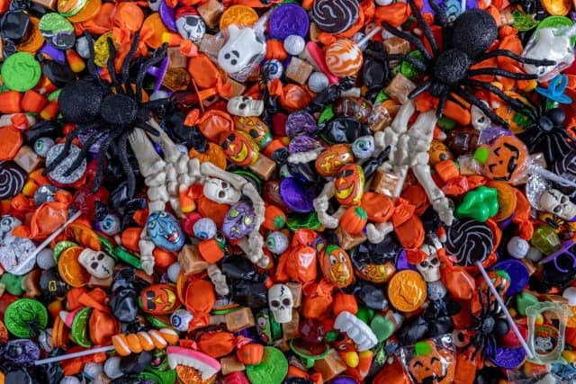 Scary Halloween skeleton hands filled with candy coming up out of a variety of Halloween candy spread out on table top and 2 big black glitter spiders