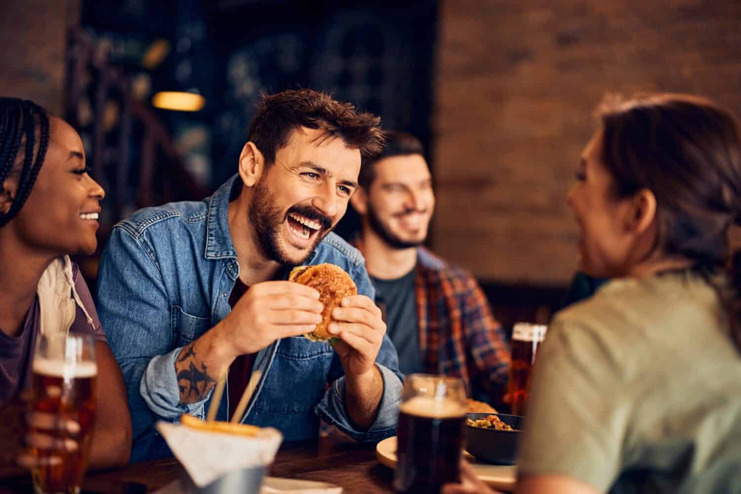 Cheerful man eating burger and having fun while gathering with friends in a bar.