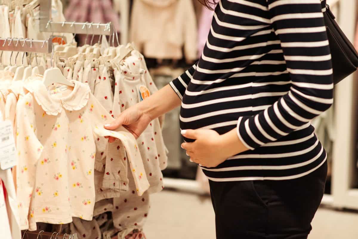 Pregnant woman choosing dress and bodysuits at clothing store. Mother doing shopping in baby shop. Baby fashion, shopping time, sale and pregnancy concept.