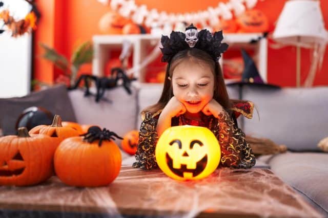 Adorable hispanic girl wearing halloween costume looking pumpkin basket at home