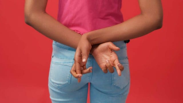 Crossed fingers. Liar sign. Fake promise. Unrecognizable dishonest woman hiding hands behind back isolated on red free space background.