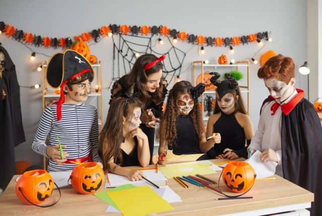 Teacher doing art and craft activity with children. Kids dressed as pirate, witch, Catrina, Maleficent and vampire having fun at Halloween party with their young English tutor at after school club