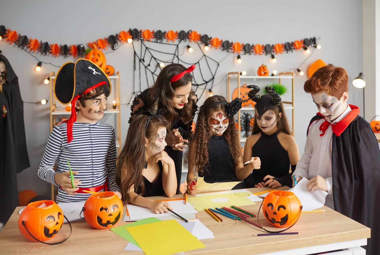 Teacher doing art and craft activity with children. Kids dressed as pirate, witch, Catrina, Maleficent and vampire having fun at Halloween party with their young English tutor at after school club