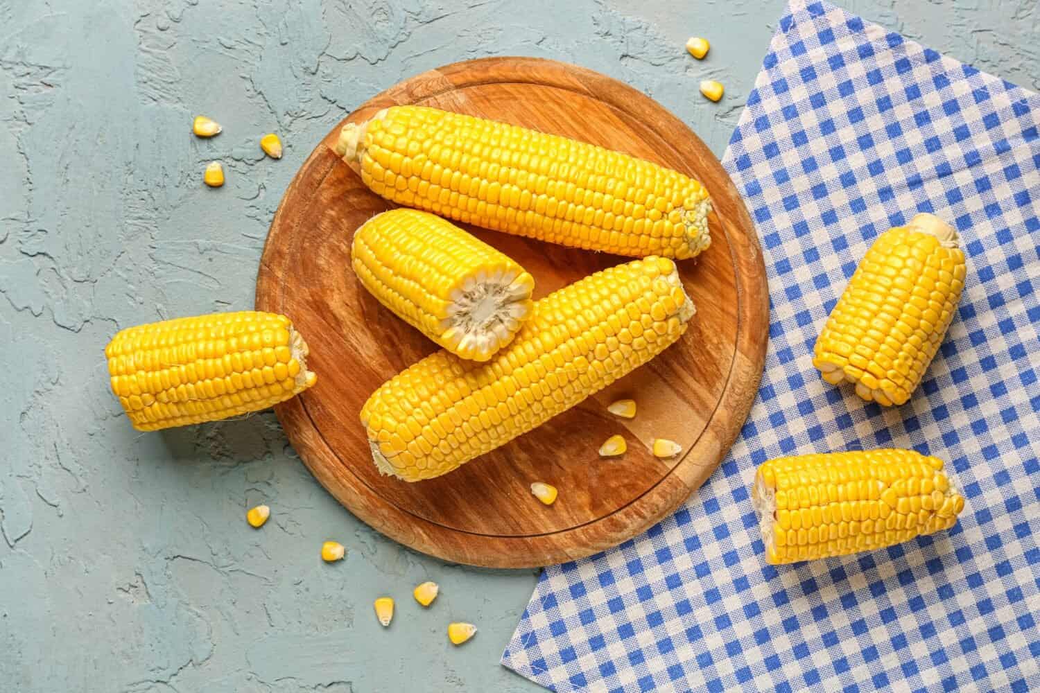 Wooden board with fresh corn cobs and seeds on blue background