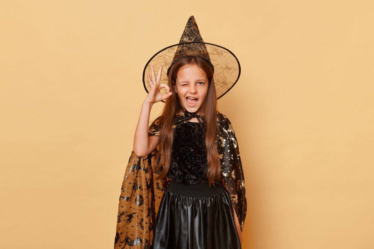 Excited little girl wearing witch costume and carnival cone hat isolated over beige background winking to camera showing okay gesture with hand.