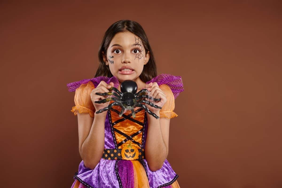 spooky girl in Halloween costume holding fake spider and grimacing on brown backdrop, October 31