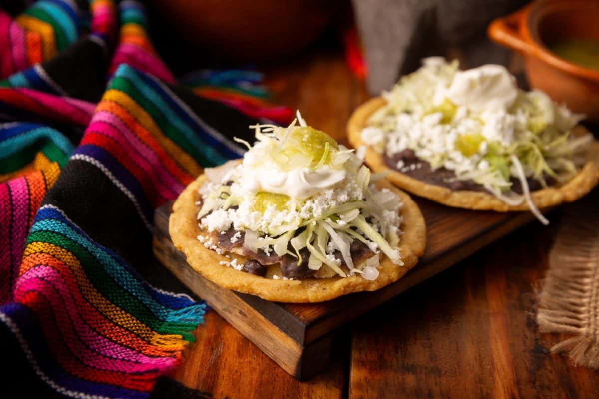 Sopes. Traditional homemade Mexican food prepared with flattened and pinched on the border fried corn dough covered with refried beans, green or red sauce, lettuce, cheese, onion and sour cream.