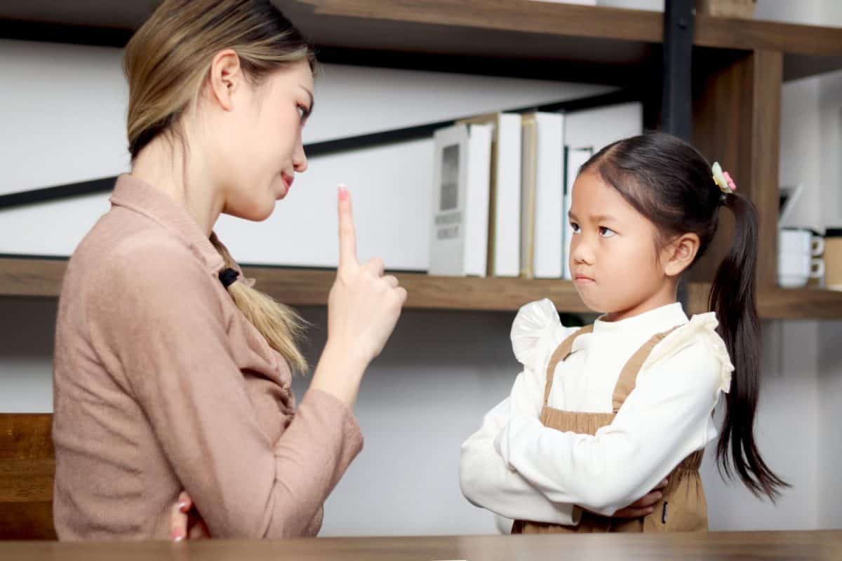 Asian unhappy angry little daughter standing with arms crossed while mother scolding her. Mom scolding child girl, teaching kid about inappropriate behavior, family relationships with conflict.