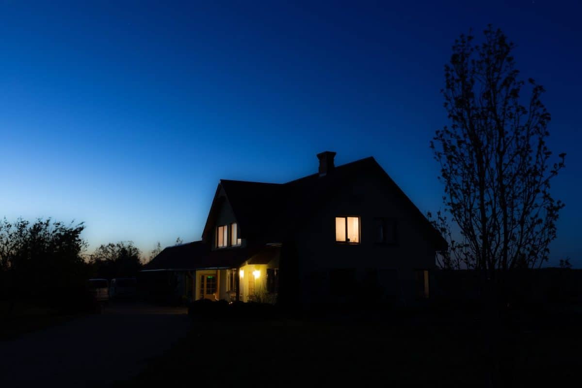 Warm lights glow from the windows of a house under the twilight sky, casting a serene ambiance over the tranquil rural setting.