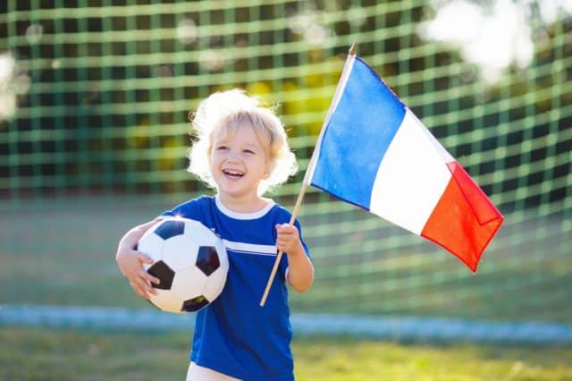 Kids play football on outdoor field. France team fans with national flag. Children score a goal at soccer game. Child in French jersey and cleats kicking ball. Fan celebrating victory at pitch.