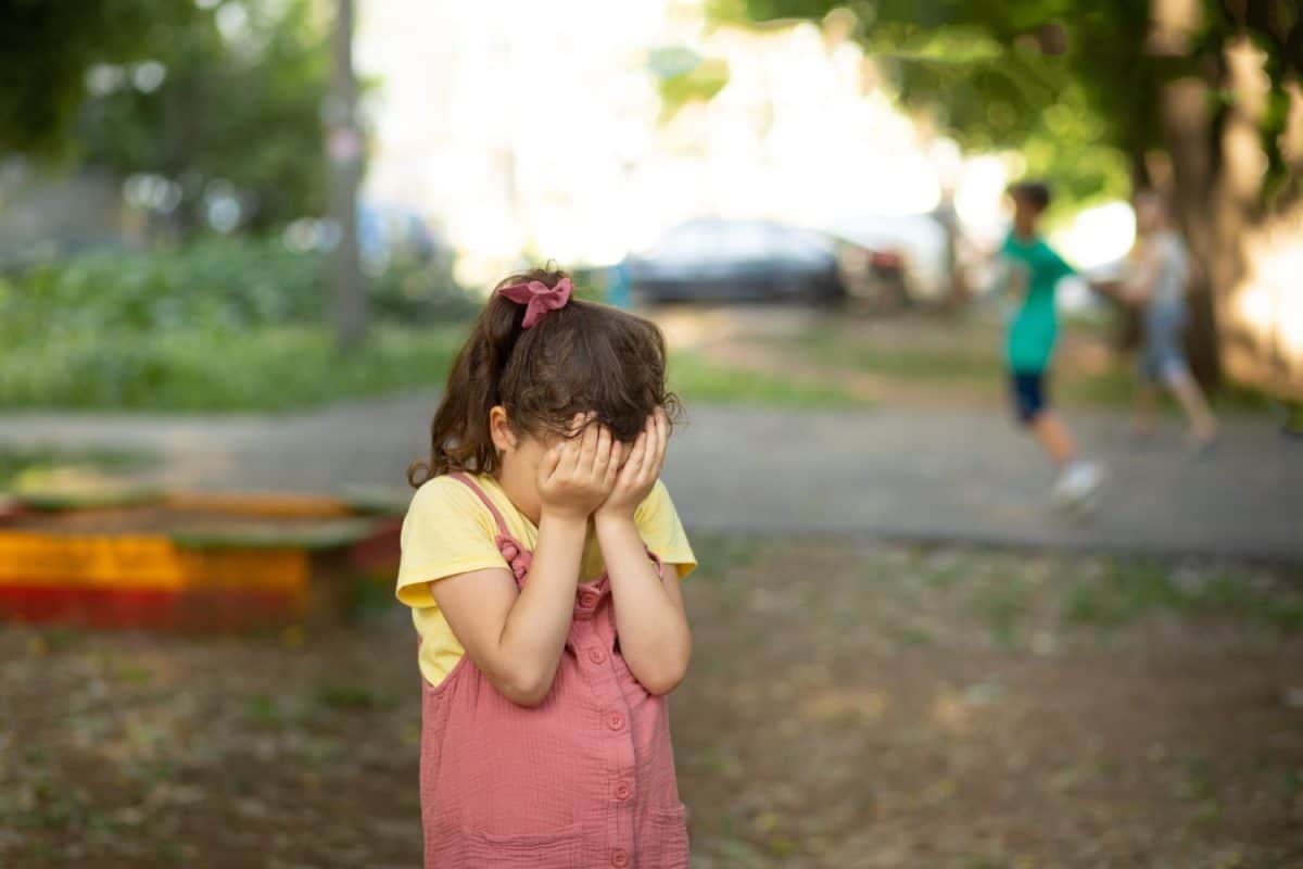 Upset lonely bullied little kid girl cries covering his eyes with his hands feels abandoned abused, sad alone preschool child in kindergarten or playground.