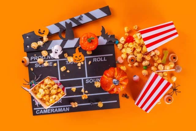 Horror movie, series night, Halloween cinema party with trick-or-treating sweets bucket, popcorn, movie clapperboard and Halloween decorations on high-colored background copy space