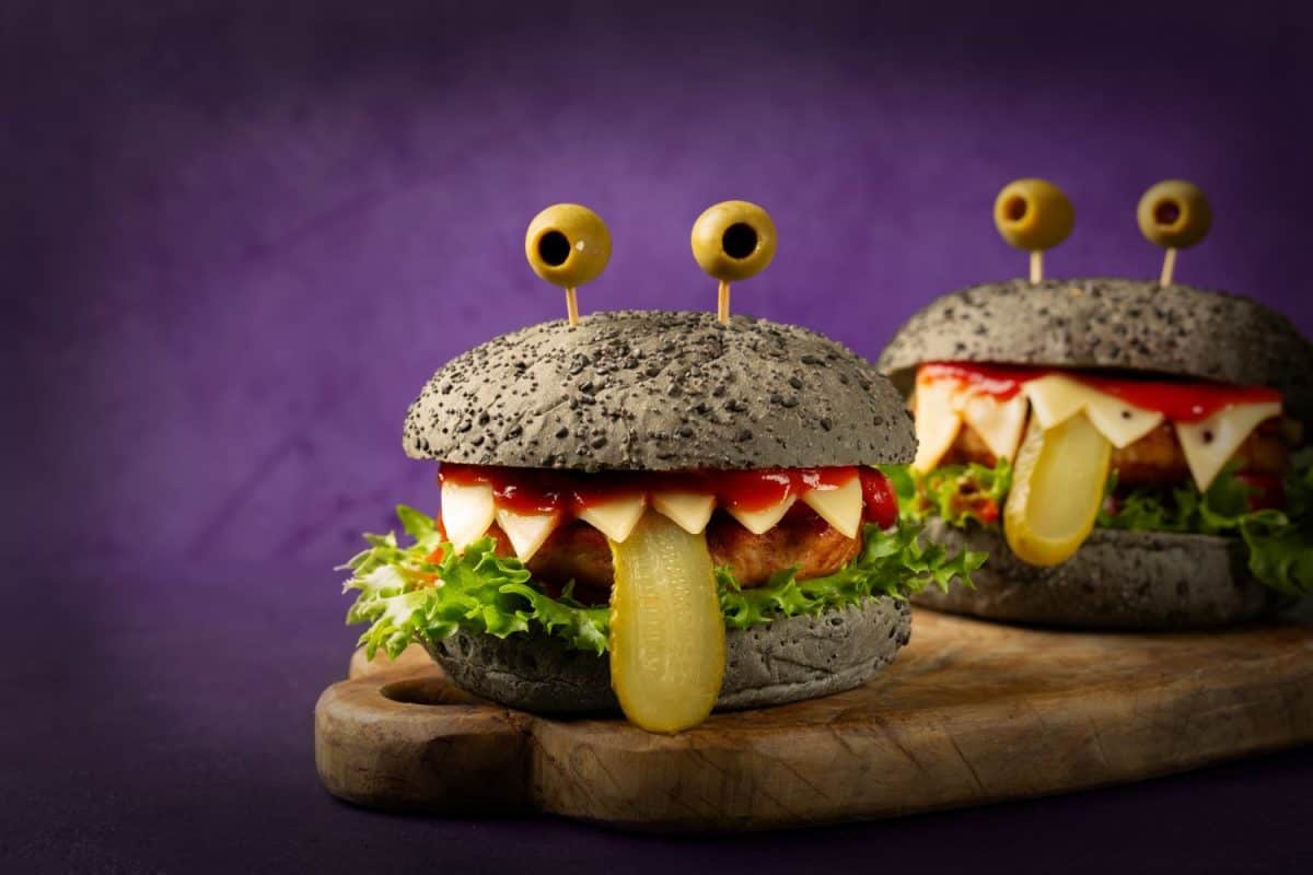 Monster burgers, fun Halloween appetizers, perfect food idea for Halloween party. Made with black charcoal buns, cheese, chicken burger, cheese, letttuce, tomato sauce. Purple background.