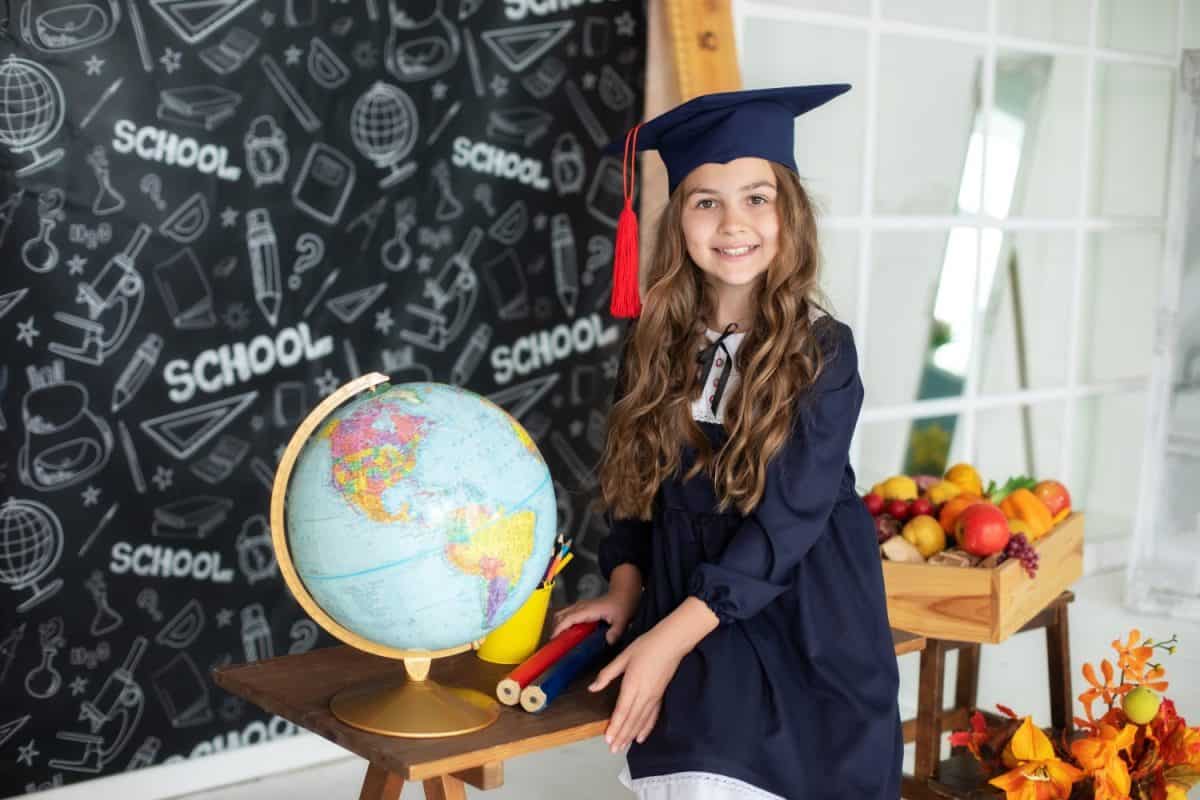 School Kid in graduate headdress is studying in classroom against of blackboard. Smiling little girl in a school uniform and master's hat holding globe in classroom. Back to school, learning lessons.