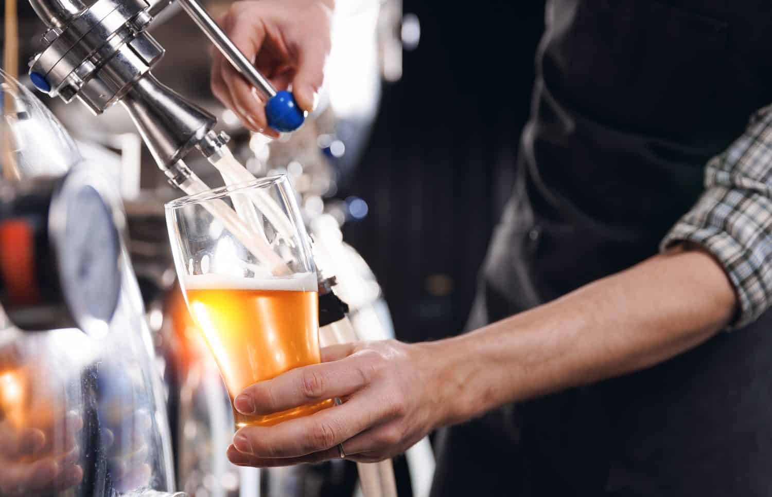 Closeup brewer pours fresh beer from steel tanker at production brewery.