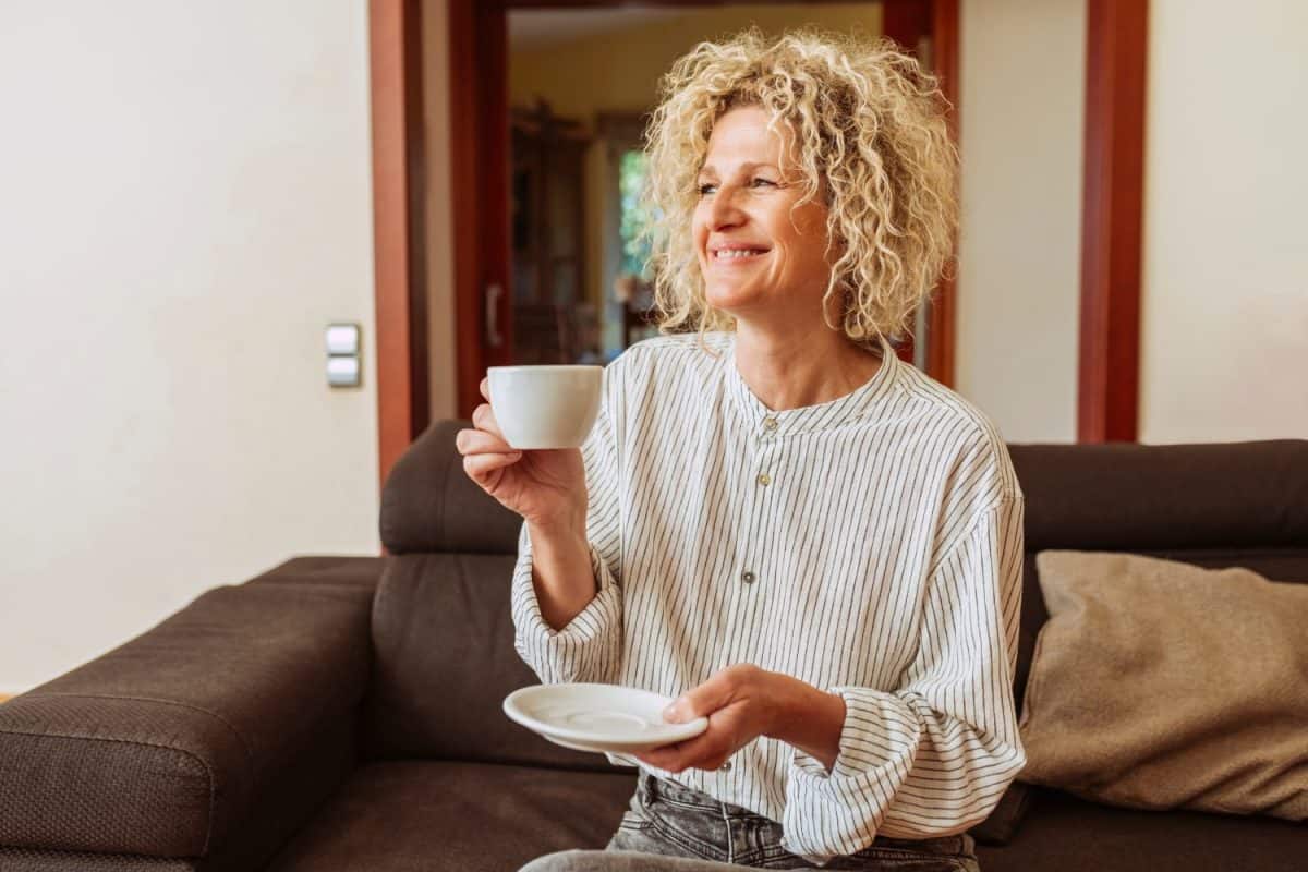 Smiling attractive relaxed middle aged female, drinks coffee sitting on couch at home. Stylish 50s lady with curly hair, enjoys resting sitting on sofa in modern living room.