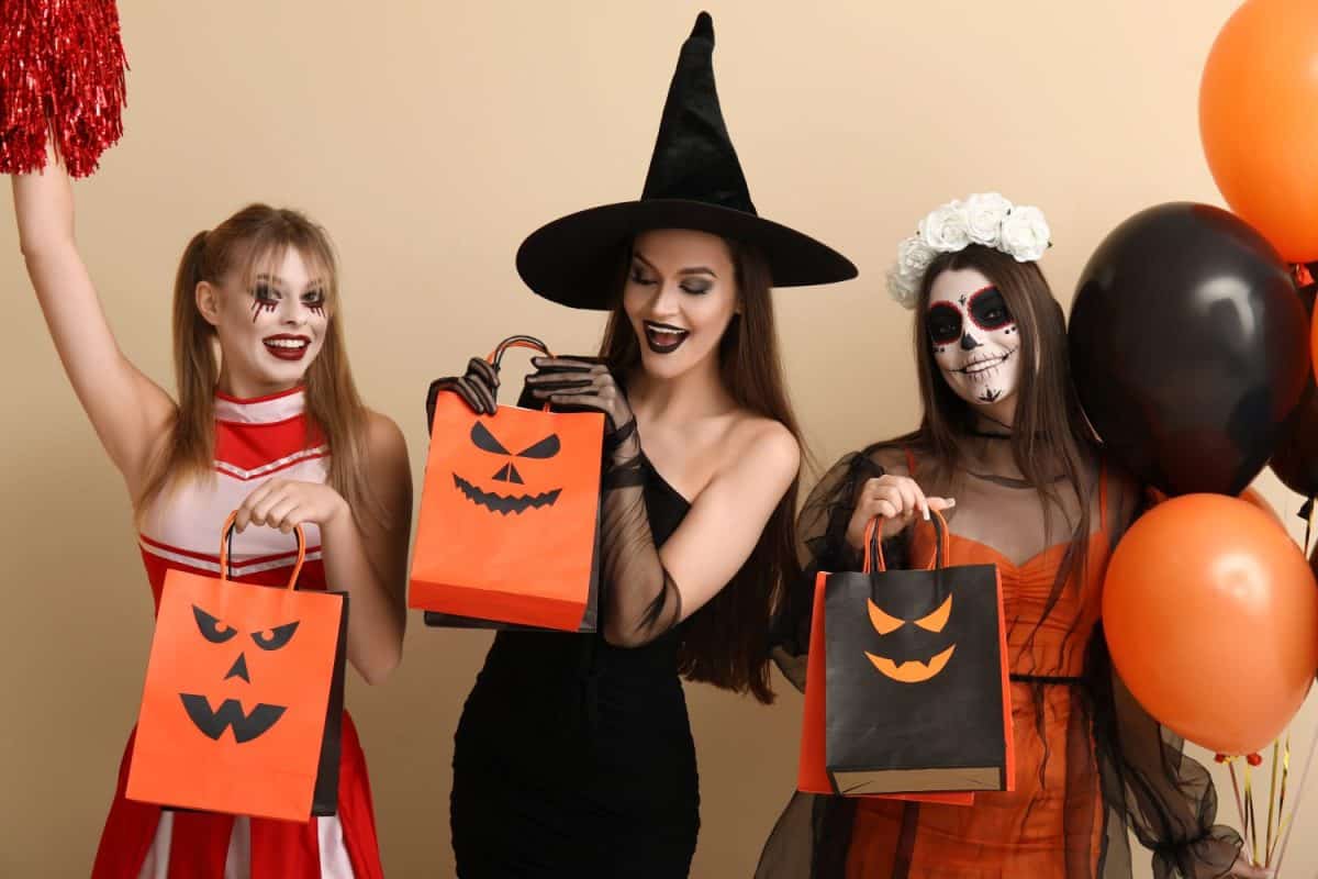 Female friends dressed for Halloween with gift bags and balloons on beige background