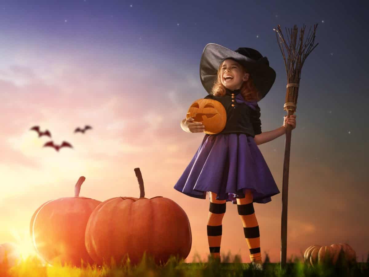 Happy Halloween! Cute little witch with a big pumpkin. Beautiful young child girl in witch costume outdoors.