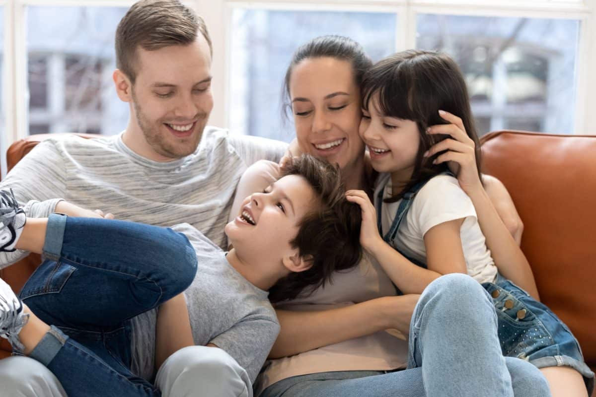 Happy mother and father hugging two children enjoying and having fun. Smiling family looking at each other. Little cute daughter and son play with mom and dad on couch.