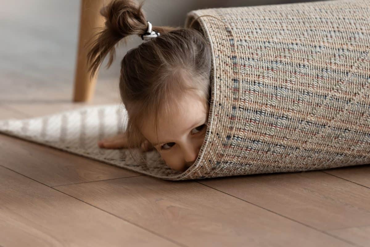 Adorable small girl wrapped in rug lying on warm wooden laminate floor in modern living room alone, close up view. Playtime funny hiding games, professional cleaning carpet company services ad concept